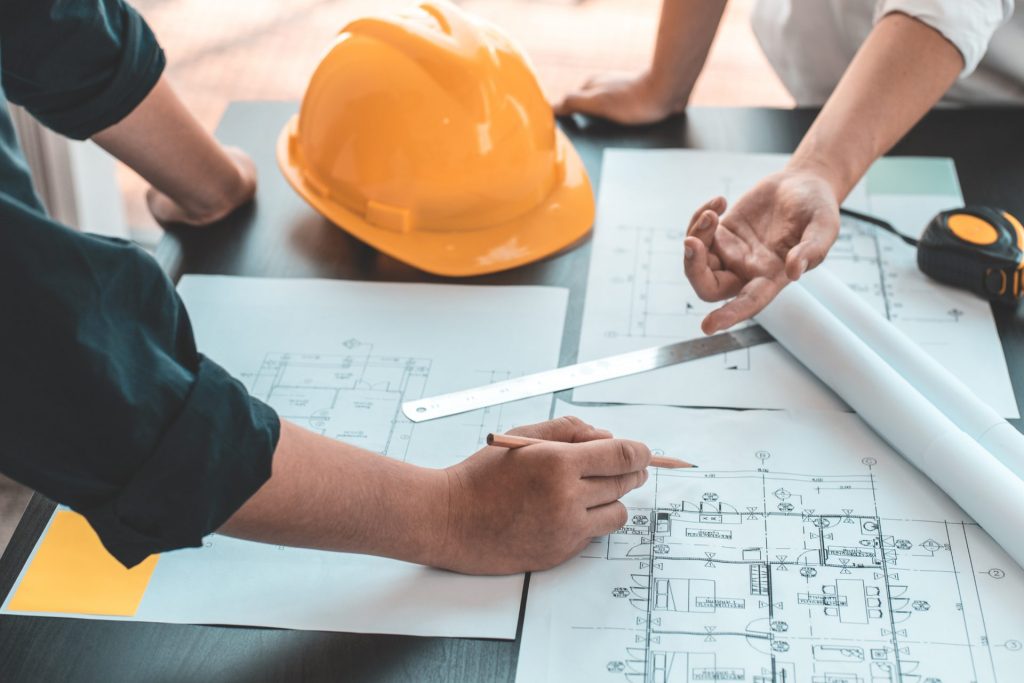 Hands reviewing building plans with a hard hat and tape measure on the desk